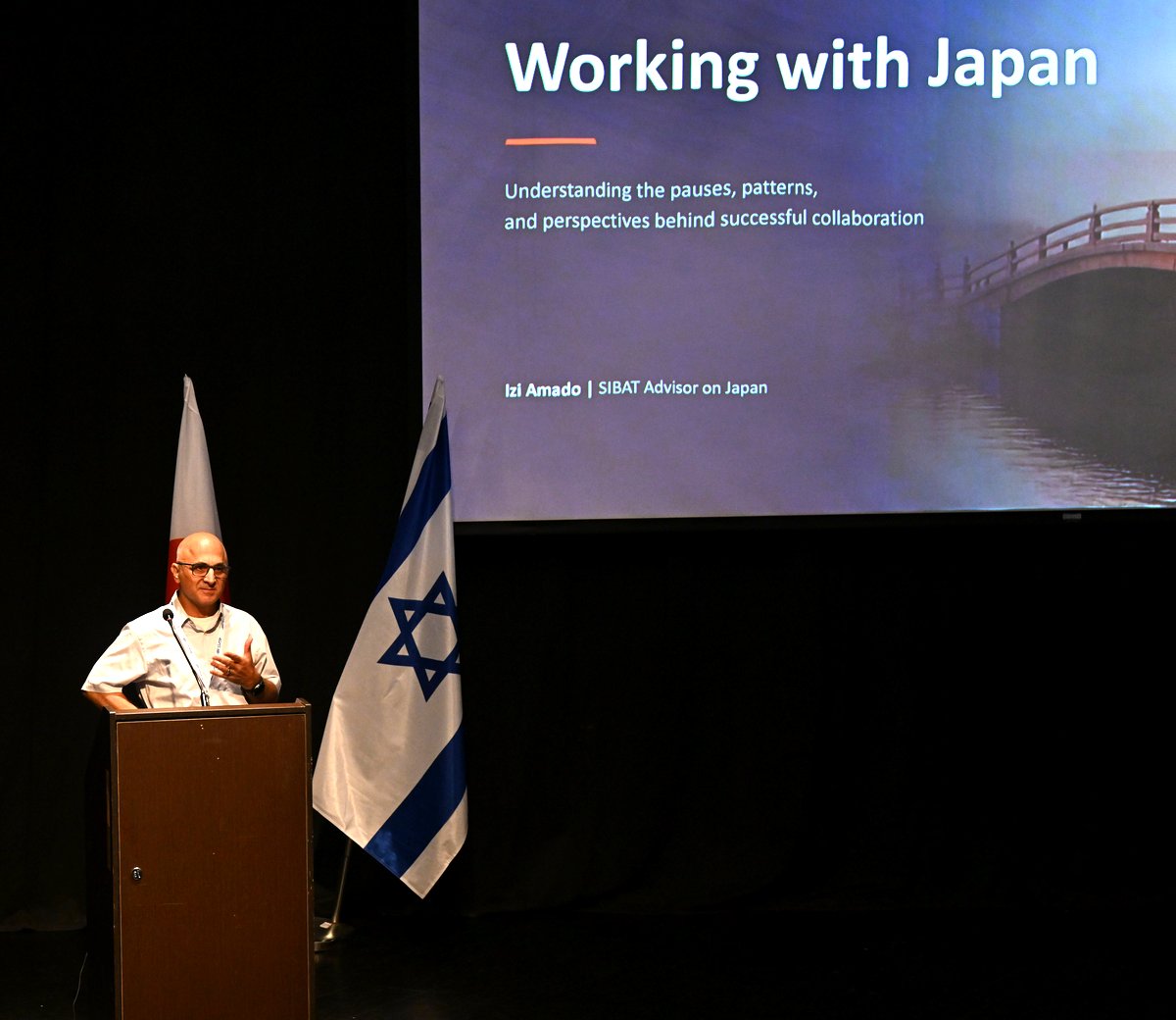 Izi Amado speaking at the Japan Seminar for SIBAT, September 2026, next to the Japanese and Israeli flags